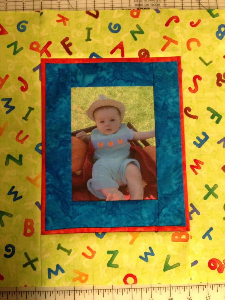 A quilt block showcasing a printed baby photo surrounded by a vibrant blue and red fabric border, with a playful alphabet-themed fabric framing the block. This custom piece is designed to be part of a larger quilt or as a unique keepsake.