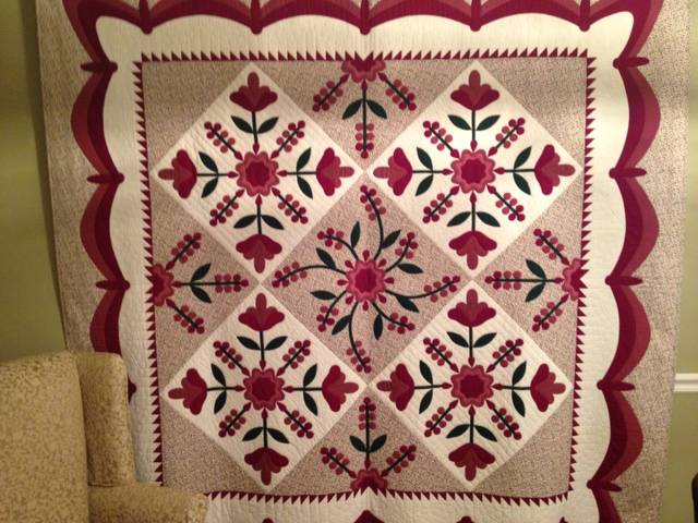 An applique quilt titled 'Hot Fudge Sundae' featuring intricate floral patterns in red and green on a cream background. Designed by Irma Gail Hatcher, the quilt is adorned with scalloped edges and detailed applique work.