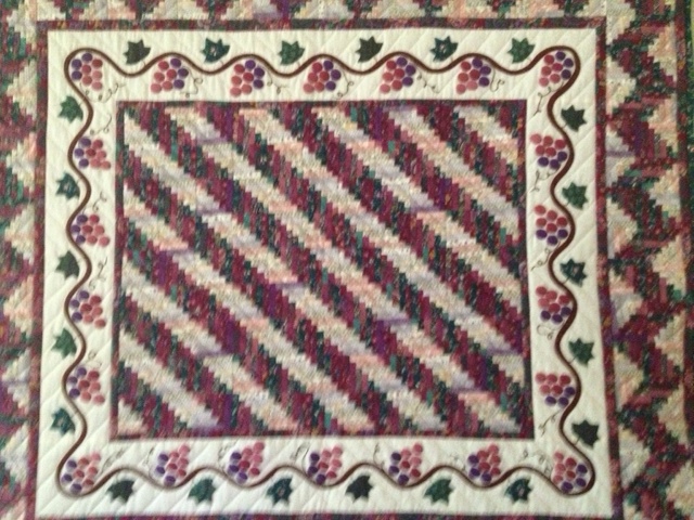 An applique quilt titled 'Log Cabin Quilt' featuring over 3,000 pieces of fabric, the smallest measuring just 3/8" x 3/8". The quilt has a diagonal striped pattern in earthy tones, framed by a grapevine border with clusters of grapes and leaves.