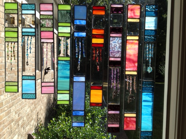 A series of narrow, rectangular stained glass panels hanging in a window, illuminated by sunlight. The panels display a range of vibrant colors and patterns, with some featuring delicate beaded accents that dangle from the glass, creating a captivating play of light and color.