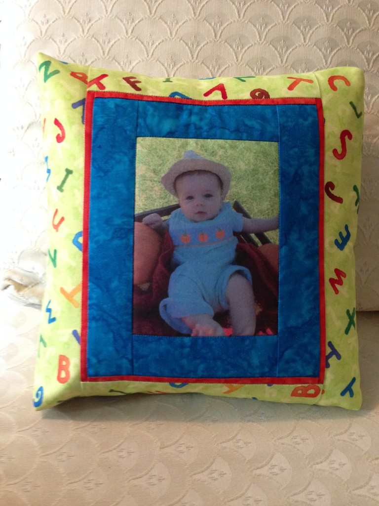 A quilt block showcasing a printed baby photo surrounded by a vibrant blue and red fabric border, with a playful alphabet-themed fabric framing the block. This custom piece is designed to be part of a larger quilt or as a unique keepsake.