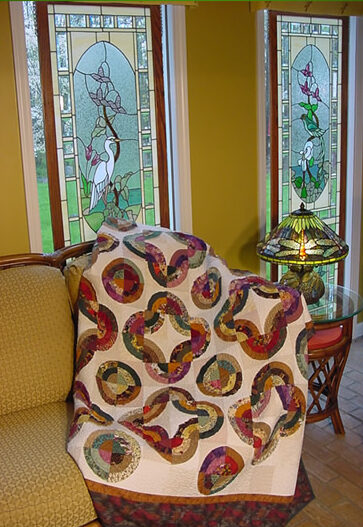 An inviting interior scene featuring a colorful quilt with intricate circular patterns draped over a yellow couch. The quilt is displayed in front of two tall stained glass windows, each adorned with floral designs adding to the cozy, artistic ambiance.