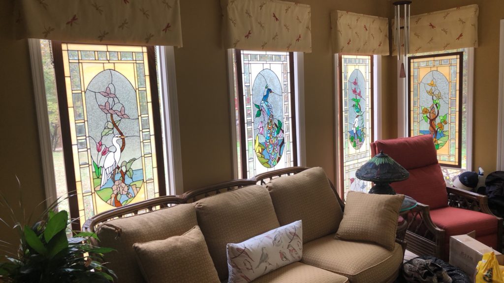 A set of four stunning 6-foot stained glass windows, each featuring intricate floral and nature designs based on Judy Miller patterns. The windows are beautifully illuminated by natural light, adding a vibrant and artistic ambiance to the cozy sitting area with a plush couch and an accent chair.