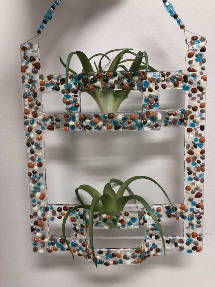 A fused glass air plant holder with a geometric frame decorated with colorful glass pebbles in shades of blue, brown, and white, holding two air plants.