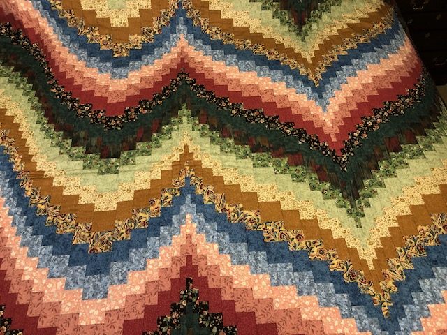 A vibrant Bargello quilt with a zigzag chevron pattern in rich shades of green, blue, red, and gold. The intricate design creates a wave-like effect, highlighting the precise piecing and color transitions.