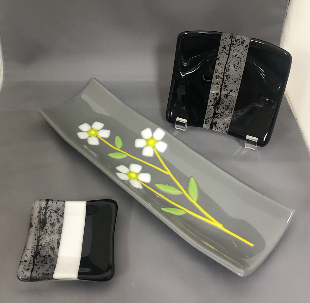 A set of fused glass dishes in black and grey tones, featuring a long platter with white flowers, a small square dish with a black and white stripe, and a black dish with a speckled grey accent.