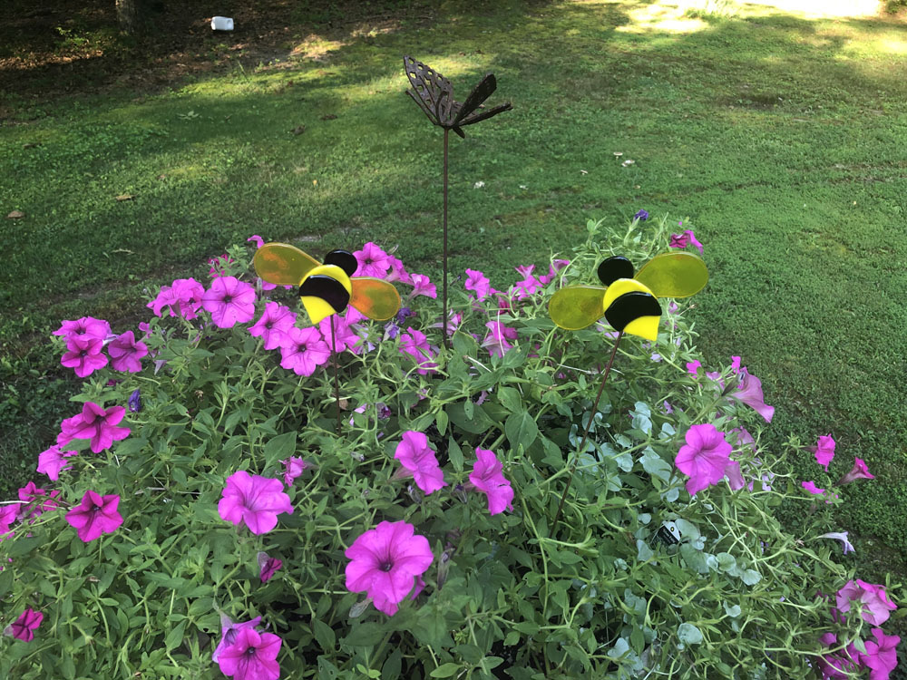 Two fused glass bumblebee plant stakes with yellow and black striped bodies, placed among vibrant purple flowers in a garden setting.