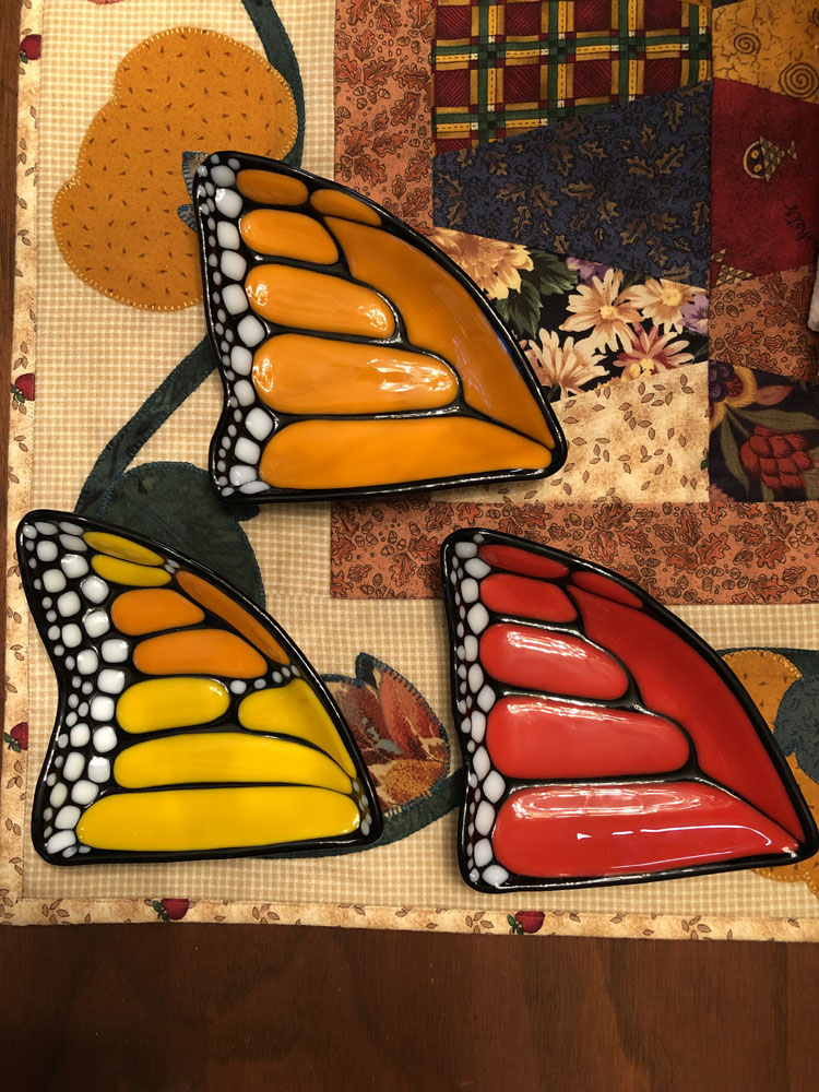 Fused glass dishes shaped like vibrant butterfly wings, available in orange, yellow, and red, each featuring detailed vein patterns and white spots along the edge.