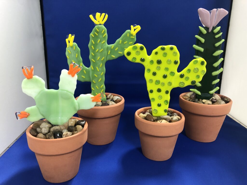 A set of four fused glass cactus sculptures, each with unique designs and colorful blooms, set in small terracotta pots filled with decorative stones.