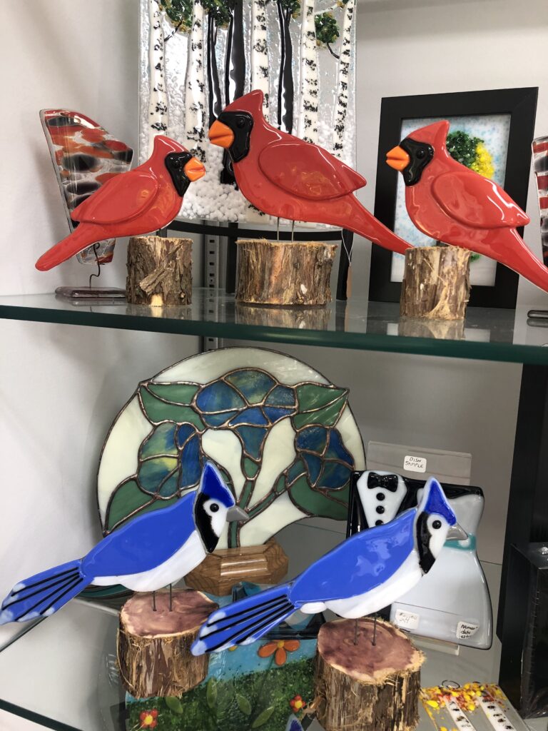 A display of fused glass cardinals and bluebirds, each perched on cedar stands. The cardinals are vivid red with black masks, while the bluebirds are cobalt blue with white chests.