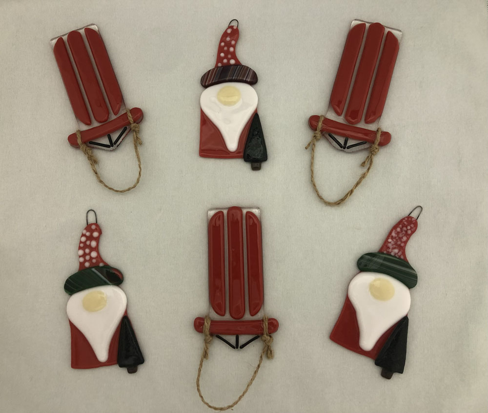 Six fused glass Christmas ornaments arranged on a light background, including three red sleds with jute rope handles and three gnomes with pointed hats, white beards, and colorful attire. The gnomes and sleds have a whimsical, handcrafted design.