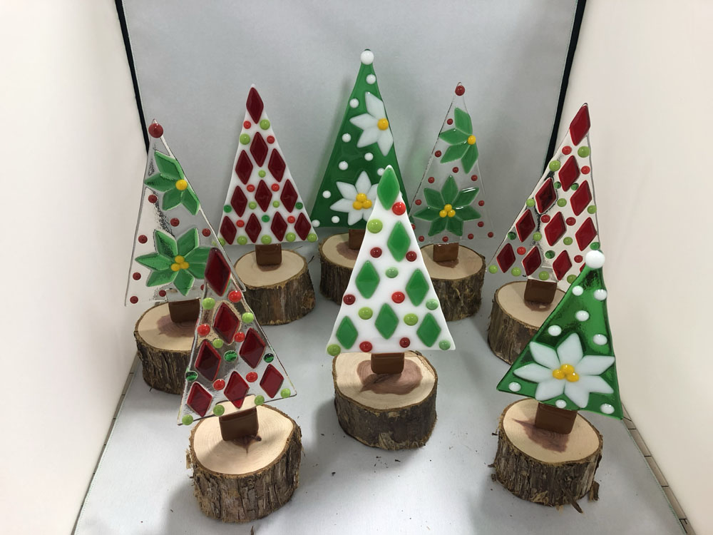 Eight handcrafted fused glass Christmas tree ornaments in festive red, green, and white designs. Each tree is mounted on a natural cedar wood base, cut from branches sourced from the artist's property. The trees feature various patterns, including poinsettias, polka dots, and diamond shapes, creating a charming holiday display.