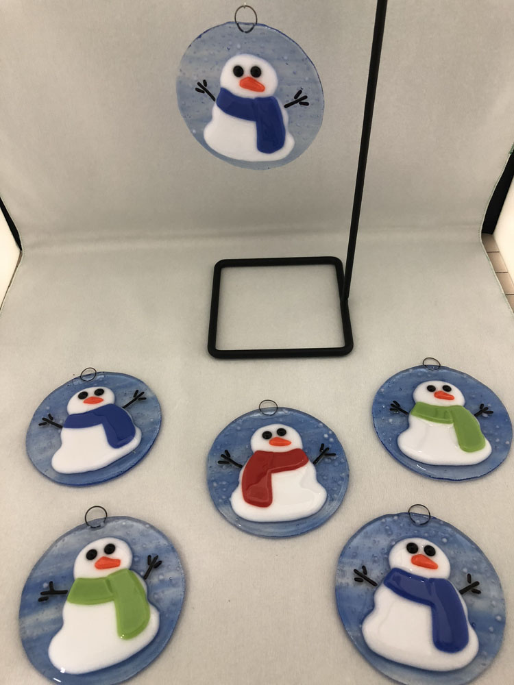 A set of circular fused glass ornaments featuring a cheerful snowman design on a blue background. The snowmen have carrot noses, black eyes, and colorful scarves in red, green, and blue.