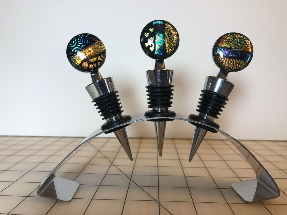 A set of three dichroic glass wine stoppers with shimmering, iridescent designs, displayed on a sleek metal holder.