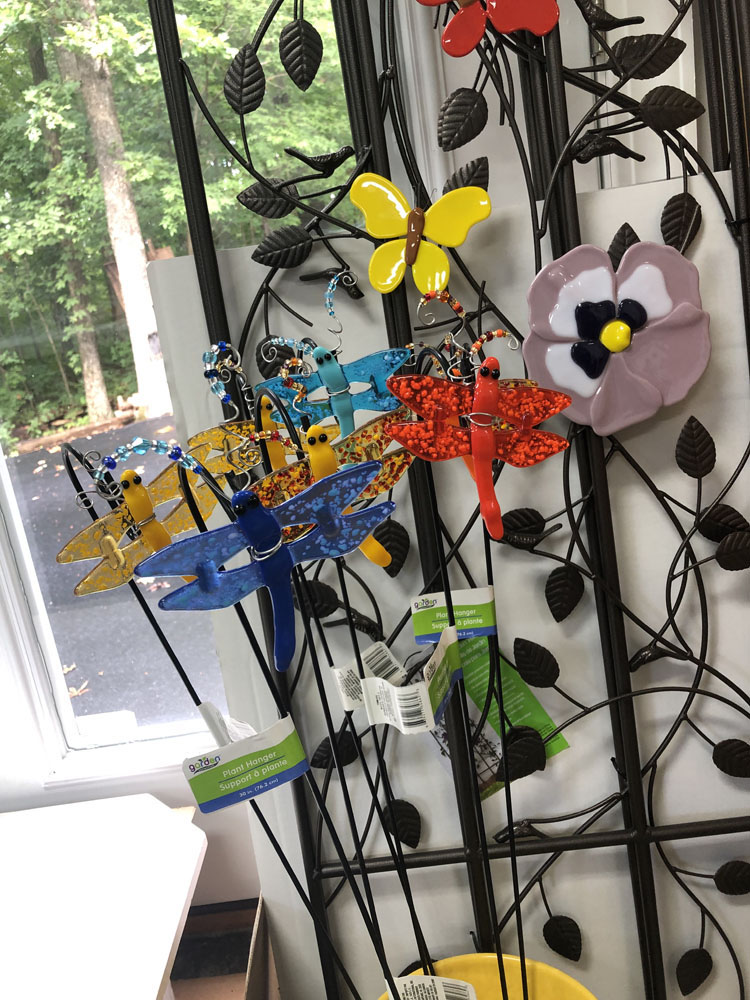 A collection of fused glass dragonfly plant stakes in various colors, along with a yellow butterfly and a purple pansy flower, displayed on a decorative metal trellis.