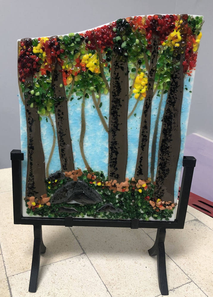 A fused glass panel depicting a forest scene with tall trees adorned with colorful autumn leaves in shades of red, orange, and yellow against a blue sky.