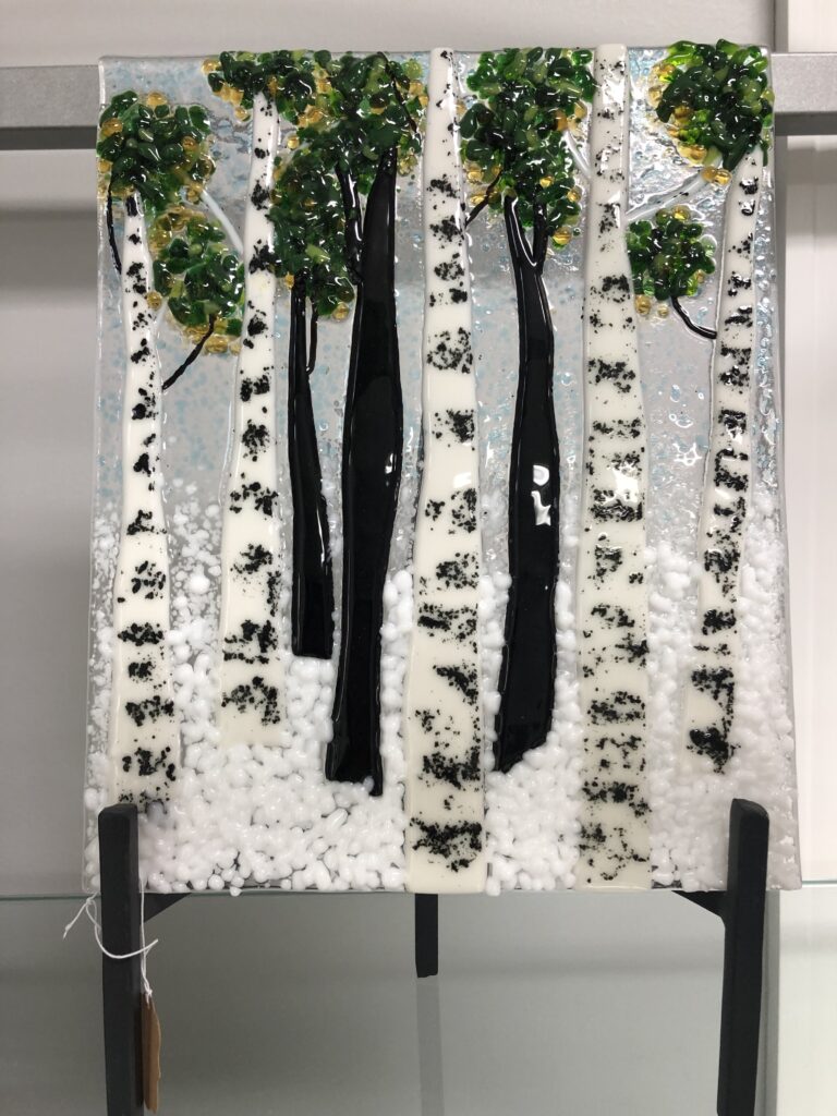 A fused glass panel featuring tall aspen trees dusted with snow, with green and yellow leaves peeking through, set against a wintry sky.