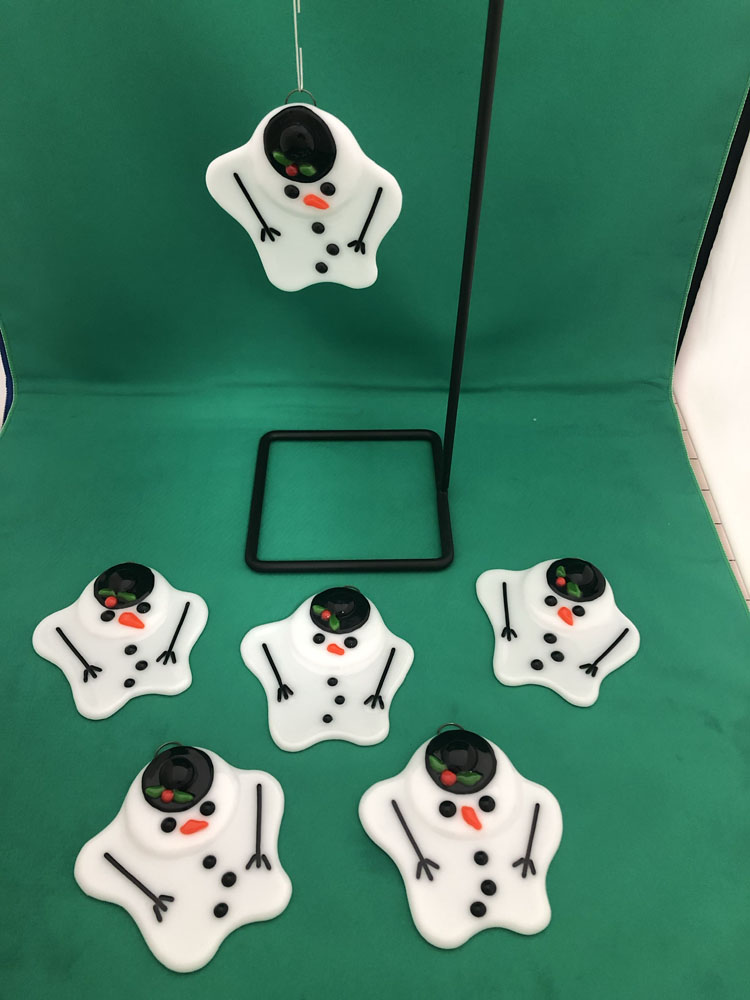 Six fused glass melting snowman ornaments displayed on a green background. The snowmen have black hats, orange carrot noses, and black buttons, with drooping arms and melting bodies. One ornament is hanging on a metal stand, showcasing its design.