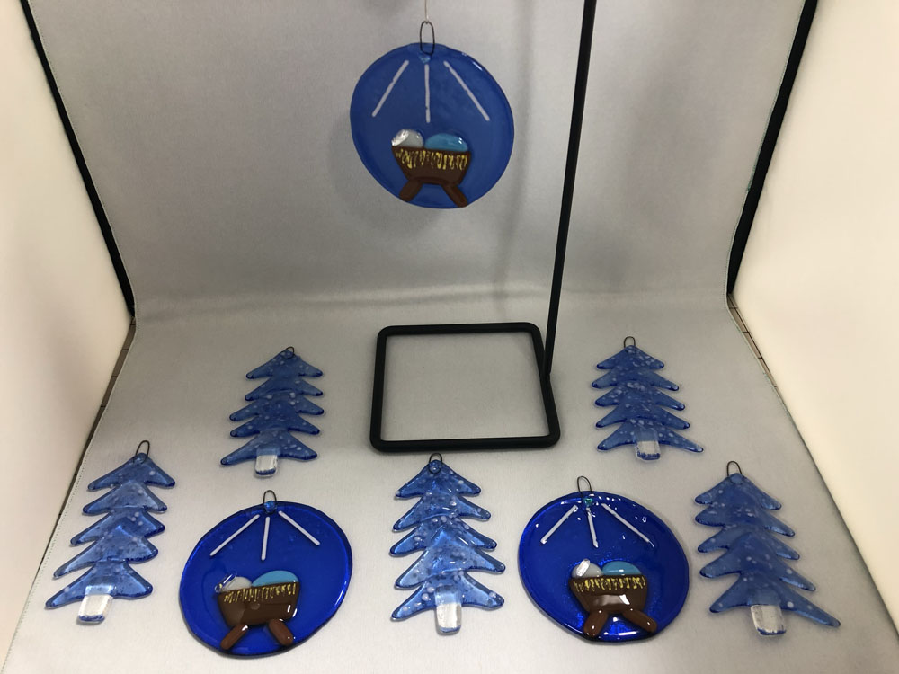 A collection of fused glass Christmas ornaments featuring blue trees and nativity scenes. The nativity ornaments showcase a baby in a manger under a star, while the tree ornaments are stylized in shades of blue with white speckles.