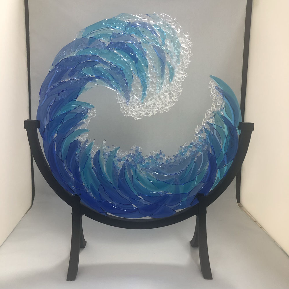 A fused glass sculpture designed to resemble an ocean wave. The wave is made from layers of blue, turquoise, aqua, teal glass, with the blue pieces forming the crest of the wave as it curls over. The ends of the curl are accentuated with clear curved glass and a spattering of tiny pieces of white and clear glass (frit). The sculpture is displayed on a black metal stand, and the texture of the glass gives the piece a dynamic, fluid appearance.