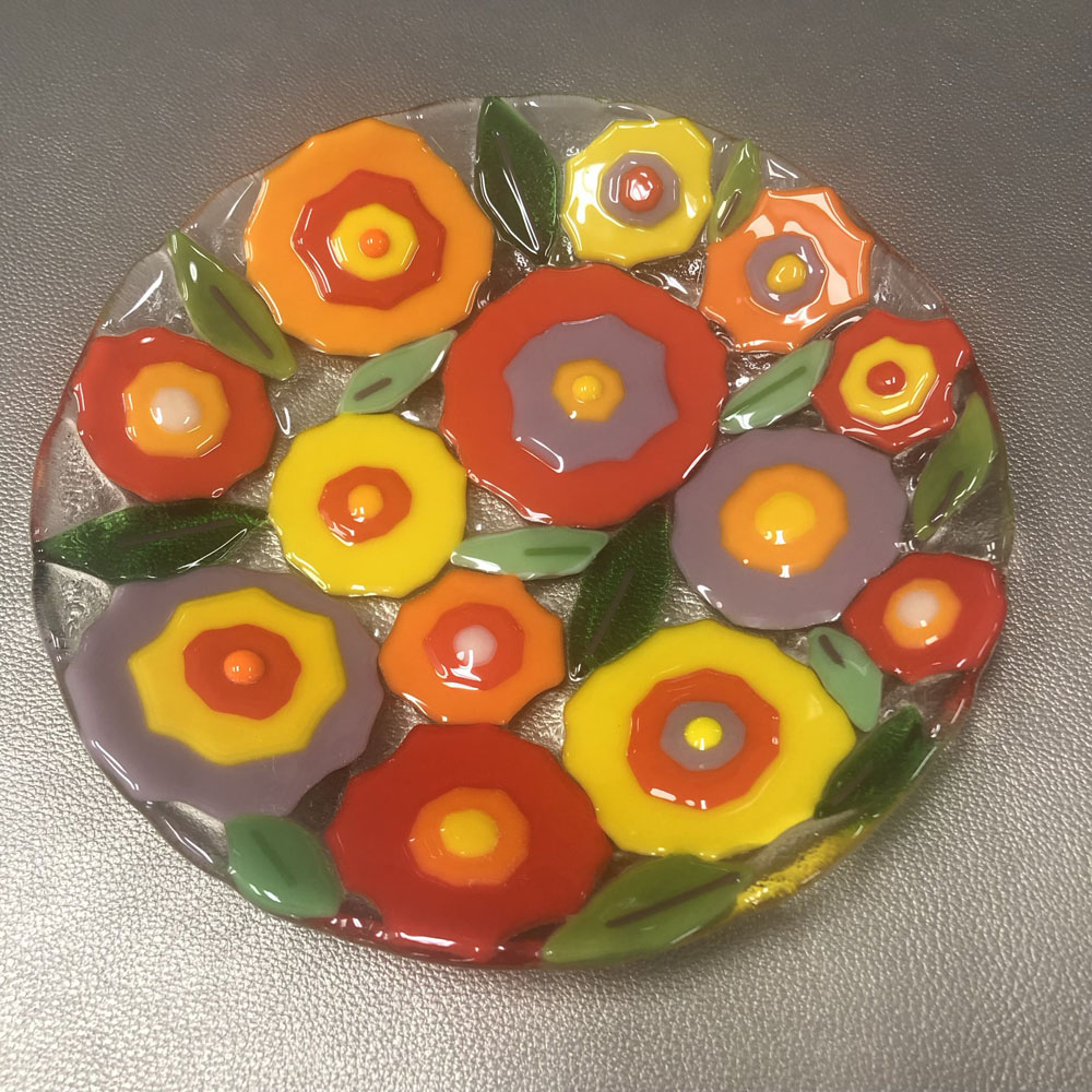 A fused glass plate decorated with colorful, abstract flowers. The flowers are arranged in a somewhat circular pattern, in a mix of bright red, orange, yellow and purple petals, with green leaves interspersed between them. The plate has a glossy surface, but you can feel the texture created by the different layers of flower petals. The vibrant colors of the flowers stand out against the clear glass background.