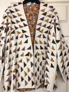 A handcrafted Prairie Point quilted jacket featuring a striking pattern of multicolored triangles on a cream background. The jacket’s unique design includes 3D points that resemble scales, adding a tactile element to the piece. The inside lining showcases a floral fabric, providing a beautiful contrast to the geometric design.