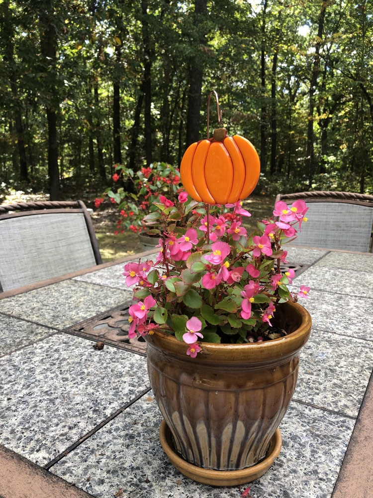 A bright orange fused glass pumpkin plant stake placed in a pot of pink flowers, set on an outdoor patio with a forested background.