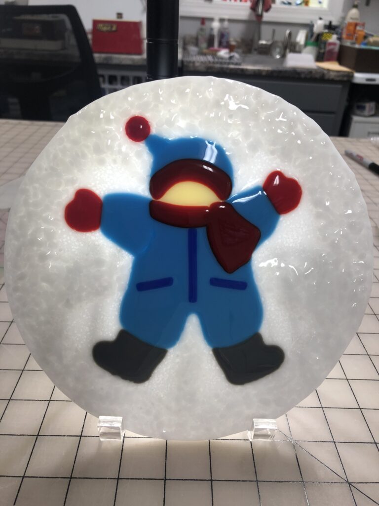 A circular fused glass artwork featuring a playful child in a blue snowsuit making a snow angel, surrounded by textured white snow.