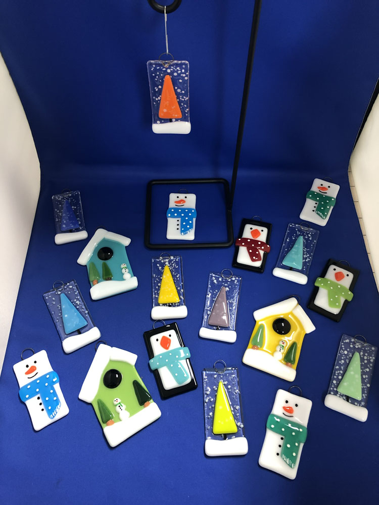 A colorful array of fused glass ornaments, including snowmen with polka dot scarves, penguins, birdhouses, and Christmas trees in various shapes and colors. The ornaments are displayed against a blue backdrop.