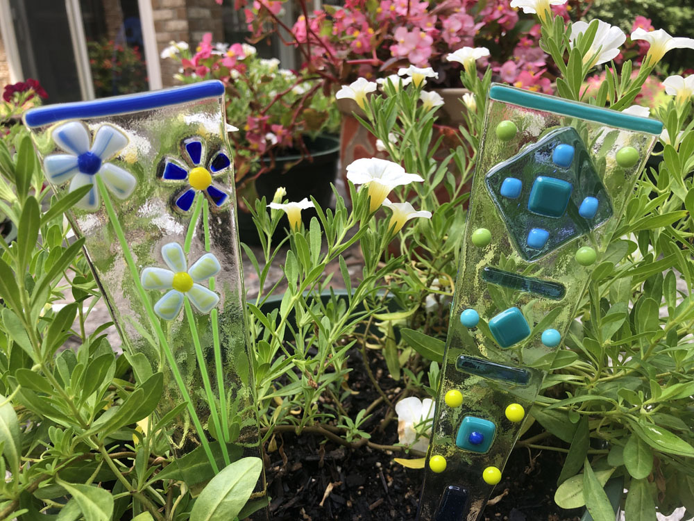 Two triangular fused glass plant stakes featuring flower designs and geometric patterns, set among lush green foliage in a garden.