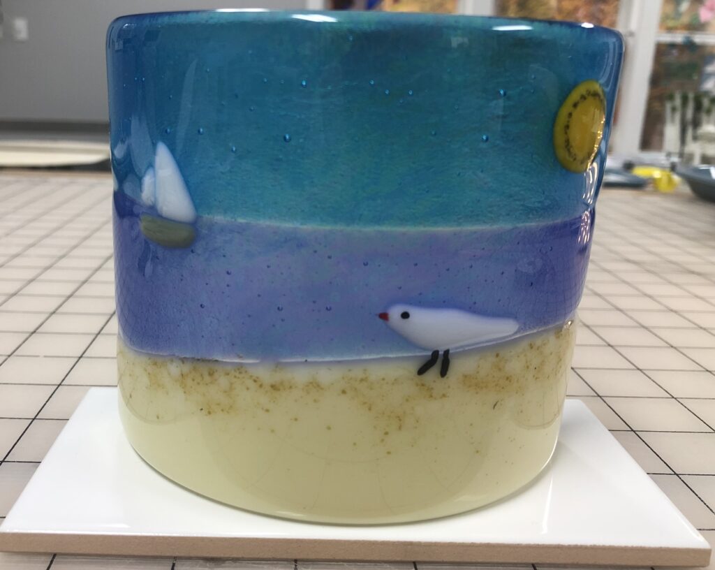 A fused glass votive candle holder depicting a serene beach scene, with a sailboat on the water, a bright sun, and a small bird on the sandy shore.