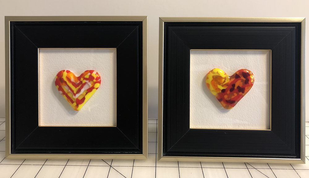 A pair of framed fused glass hearts, one with swirling patterns of red, yellow, and orange, and the other with a similar warm color palette, displayed in black frames.