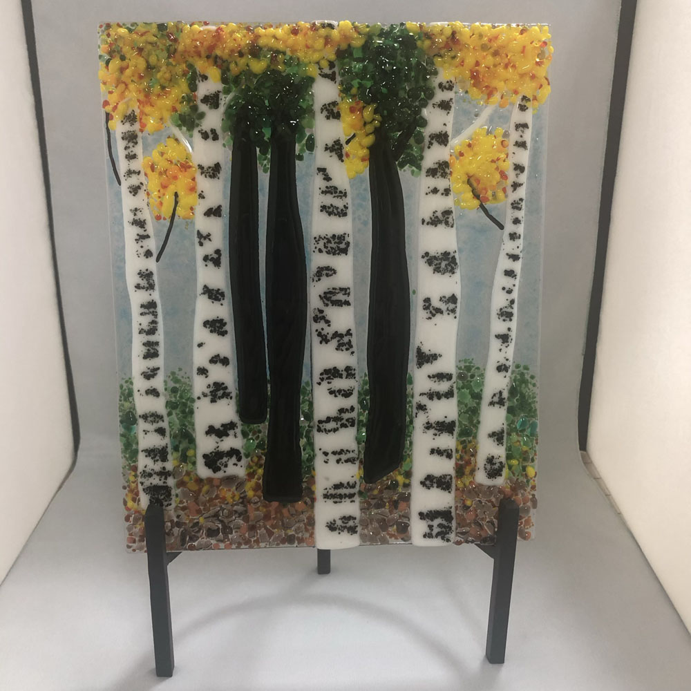 A fused glass sculpture depicting a serene forest scene with tall, slender aspen trees. The trees have white bark with black markings, set against a light blue sky background. The tree leaves are in vibrant shades of yellow, green, and orange, reflecting a forest in late summer or early autumn. The piece is displayed on a black metal stand.