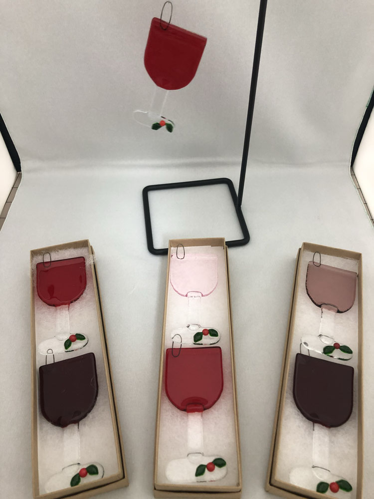 Fused glass ornaments shaped like wine glasses in shades of red, pink, and burgundy, with small holly leaf accents at the base of the glass.