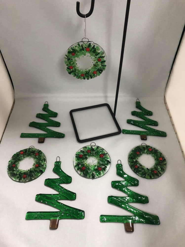 A collection of fused glass ornaments featuring green wreaths with red berries and zigzag-shaped Christmas trees. The ornaments are arranged together with one wreath displayed on a stand.